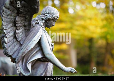 Eine geflügelte Engelsfigur im Profil auf einem herbstlichen Friedhof schaut traurig zu Boden mit einer ausgestreckten Hand Stockfoto