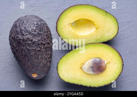 Drei Avocados, eine ganze und zwei Hälften, sind auf einer grauen Schieferoberfläche angeordnet. Die halbierten Avocados zeigen ihr cremiges gelbes Fleisch und ihr großes braunes p Stockfoto