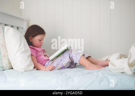 Ein junges Mädchen, das im Schlafanzug auf ihrem Bett sitzt und ein Buch liest, in einem gemütlichen, sanft beleuchteten Schlafzimmer Stockfoto