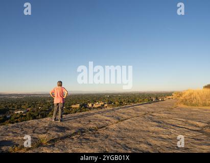 Ältere Frau mit rosa Hemd und grauer Hose auf einem Felskamm mit Blick auf eine Stadt. Fotografiert mit dem Rücken zur Kamera in Billings, Mo Stockfoto