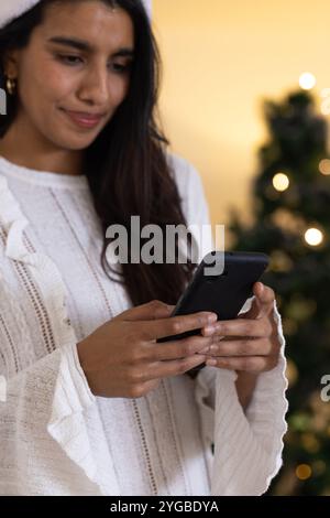 Asiatische Frau im Weihnachtsmann-Hut, die auf dem Smartphone in der Nähe des Weihnachtsbaums schreibt, zu Hause Stockfoto