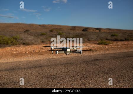 DJI-Drohne im Rahmen, schwebt über einer versiegelten Straße, kurz vor der Landung. Der australische Outback-Hintergrund ist verschwommen und zeigt roten Schmutz, Bus Stockfoto