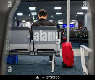 Rückansicht eines Mannes, der im Abflugbereich des Flughafens wartet. Nicht erkennbare Passanten spiegeln sich auf den Glasfenstern. Stockfoto