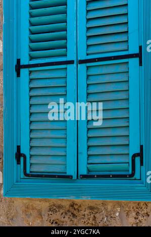 Farbenfrohe blaue Fensterläden umrahmen eine strukturierte Steinmauer, die die bezaubernde mediterrane Architektur bei warmem Sonnenlicht zeigt. Stockfoto