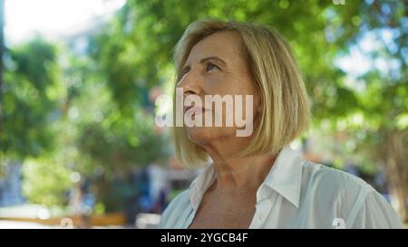 Eine Frau mit blonden, kurzen Haaren in den 50ern steht in einem urbanen Park im Freien, in einem weißen Hemd, mit Sonnenlicht, das durch grüne Bäume gefiltert wird Stockfoto