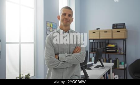 Junger Mann, der mit überkreuzten Armen in einer modernen Büroumgebung steht, mit organisierten Regalen und Büromaterialien, die in natürlichem Innenlicht erfasst werden Stockfoto