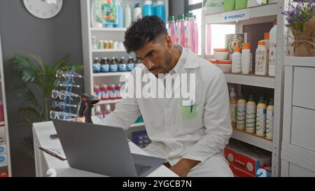 Junger hispanischer Mann, der Laborkittel trägt, der in der Apotheke mit Laptop und Regalen von Produkten im Hintergrund arbeitet Stockfoto