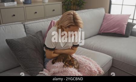 Eine junge blonde weiße Frau hält sich vor Schmerzen in den Magen, während sie in einem Wohnzimmer auf einer Couch sitzt, mit einem kleinen chihuahua-Hund auf einem rosafarbenen Rohling liegt Stockfoto