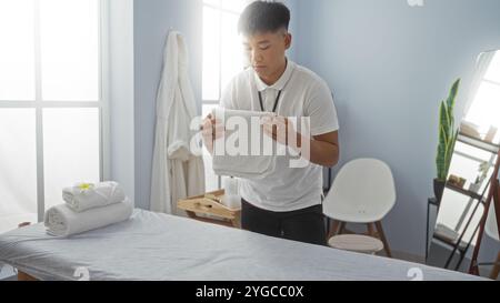 Junger chinesischer Mann in einem Wellnesscenter, der Handtücher in einem ruhigen Spa-Raum mit weißen Wänden, Massagetisch und Tageslicht durch große Fenster arrangiert. Stockfoto