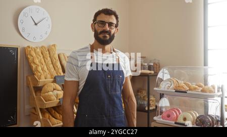 Ein hübscher junger hispanischer Mann mit Bart steht selbstbewusst in einer Bäckerei, umgeben von frisch gebackenem Brot und Gebäck. Stockfoto