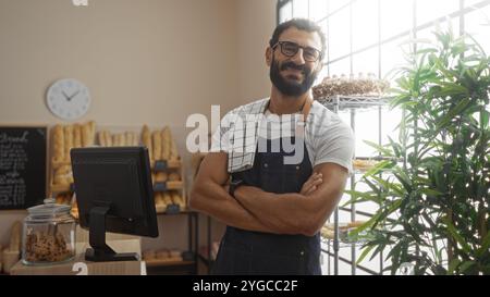 Ein lächelnder Mann mit Bart und Gläsern steht selbstbewusst mit gekreuzten Armen in einer hellen Bäckerei, umgeben von frischem Brot, Gebäck und einem einladenden atmos Stockfoto