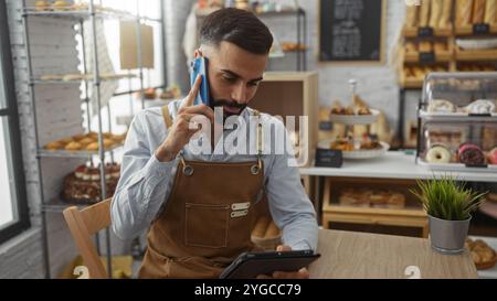 Junger Mann, der telefoniert, während er ein Tablet in einer Bäckerei benutzt, umgeben von verschiedenen Broten und Gebäck Stockfoto