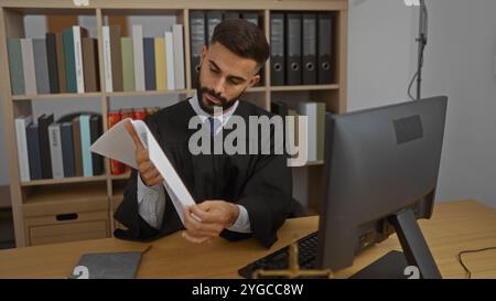 Hispanischer Mann, der in einem Büro arbeitet, in einem Richtermantel gekleidet, Dokumente an einem Schreibtisch liest, mit Computer und Bücherregal im Hintergrund. Stockfoto