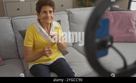 Eine ältere hispanische Frau mit kurzen Haaren sitzt in einem Wohnzimmer, mit Blick auf die Kamera, in einer komfortablen Umgebung. Stockfoto