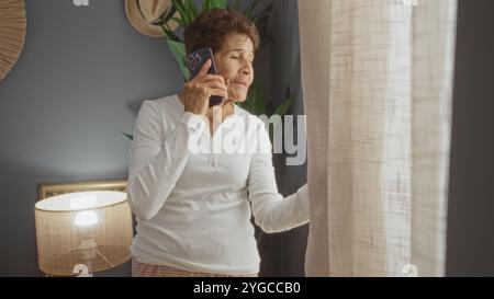 Eine ältere hispanische Frau mit kurzen Haaren steht in einem Wohnzimmer, hält ein Smartphone am Ohr und blickt zu Hause aus dem Fenster. Stockfoto