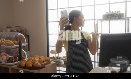 Junge Frau in einer Bäckerei, die Produkte per Video zeigt, drinnen mit Gebäck und Kuchen steht, heller Hintergrund. Stockfoto