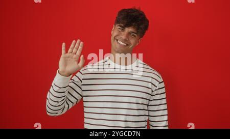 Hübscher junger hispanischer Mann, der in einem gestreiften Hemd vor einem isolierten roten Hintergrund lächelt und winkt. Stockfoto
