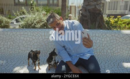 Ein hispanischer Mann mittleren Alters macht an einem sonnigen Tag ein Selfie mit zwei chihuahua Hunden auf einer gekachelten Bank in einem Stadtpark. Stockfoto