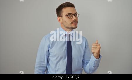 Junger hispanischer Mann mit Brille und Bart, der die Daumen in einem blauen Hemd hochlegt und vor einem isolierten weißen Hintergrund bindet. Stockfoto