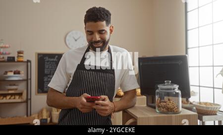 Junger Mann in einer Bäckerei mit Schürze und Lächeln, während er mit Keksen auf sein Handy in der Nähe der Theke schaut Stockfoto