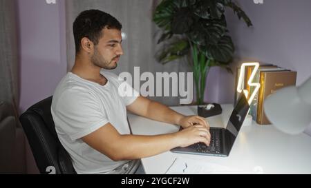Junger hispanischer Mann, der an einem Laptop arbeitet, in einem modernen Wohnzimmer mit Pflanzen und Neonlampe Stockfoto