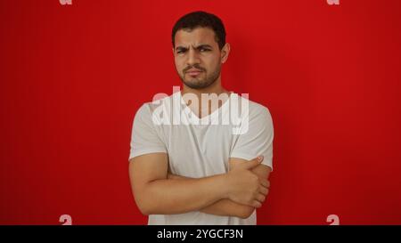 Hübscher junger hispanischer Mann, isoliert vor einem auffälligen roten Hintergrund mit einem besinnlichen Ausdruck in einem lässigen weißen T-Shirt Stockfoto