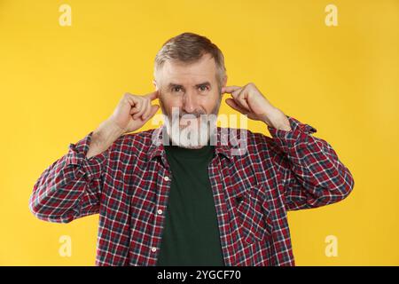 Senior-Mann, der seine Ohren mit Fingern auf orangefarbenem Hintergrund bedeckt Stockfoto