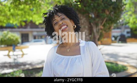 Eine wunderschöne junge afroamerikanische Frau mit lockigen Haaren lächelt hell, während sie an einem sonnigen Tag draußen in einem urbanen Park steht. Stockfoto