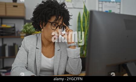 Lockhaarige afroamerikanerin mit Brille, sitzt drinnen an einem Arbeitsplatz und sieht nachdenklich aus, während sie einen Computer benutzt. Stockfoto
