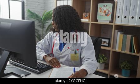 Eine junge afroamerikanische Ärztin mit lockigen Haaren arbeitet an einem Computer in einem Klinikbüro, umgeben von Bücherregalen und medizinischen Akten. Stockfoto