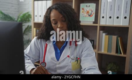 afroamerikanische Frau mit lockigem Haar in einer Klinik, mit weißem Mantel und Stethoskop, die an ihrem Schreibtisch mit medizinischen Akten im Hintergras arbeitet Stockfoto