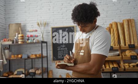 Junger Mann in der Bäckerei, der Notizen macht, umgeben von frischem Brot und Gebäck Stockfoto