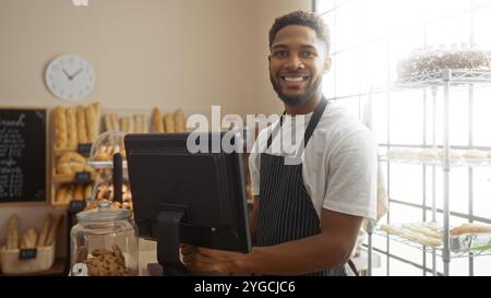 Junger Mann, der in einer Bäckerei arbeitet und die Kamera anlächelt, der hinter der Theke steht, mit Regalen mit Brot und Süßigkeiten im Hintergrund Stockfoto