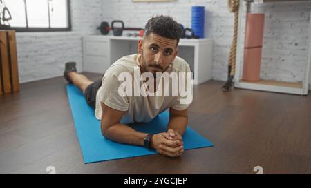 Ein hübscher junger hispanischer Mann dehnt sich bequem auf einer blauen Yogamatte in einem modernen Fitnessstudio mit verschiedenen Trainingsgeräten. Stockfoto