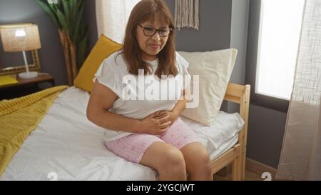 Eine ältere hispanische Frau sitzt auf einem Bett in einem gemütlichen Schlafzimmer und leidet unter Magenschmerzen. Sie reflektiert einen Moment der Unannehmlichkeit in einer heimeligen Innenumgebung. Stockfoto