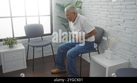 Hispanischer grauhaariger Mann mittleren Alters mit Schmerzen, sitzend in einem Wartezimmer mit weißen Wänden und grauen Stühlen, mit dem Magen umklammert und Unwohlsein vermittelt Stockfoto