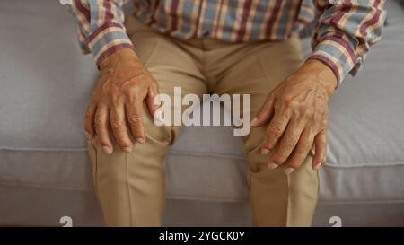 Mann mittleren Alters mit Händen, die auf Knien ruhen, sitzt drinnen auf einem Sofa in einem Wohnzimmer und trägt ein kariertes Hemd und Khaki-Hose. Stockfoto