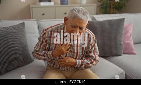 Ein älterer hispanischer Mann mit grauen Haaren hat Brustschmerzen, während er zu Hause auf einer Couch im Wohnzimmer sitzt. Stockfoto