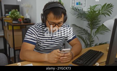 Ein junger asiatischer Mann, der Kopfhörer trägt, nutzt ein Smartphone in einer Büroumgebung mit Topfpflanzen und Computergeräten. Stockfoto