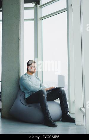Ein junger Manager in Brille und lässiger Kleidung sitzt auf einem weichen Stuhl und arbeitet an einem Laptop. Arbeitsplatz für Fernarbeit. Stockfoto