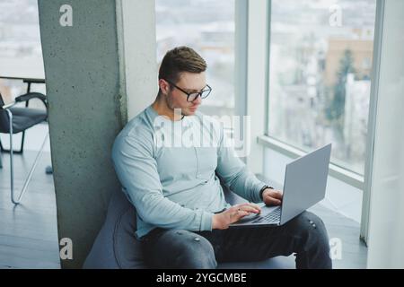 Ein junger Manager in Brille und lässiger Kleidung sitzt auf einem weichen Stuhl und arbeitet an einem Laptop. Arbeitsplatz für Fernarbeit. Stockfoto