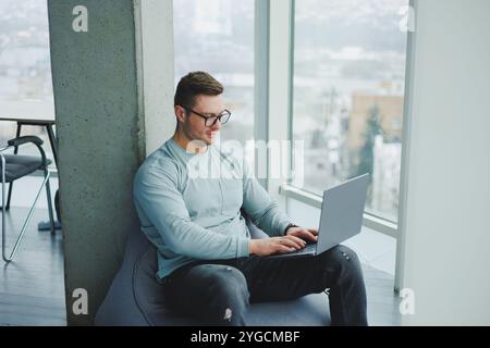 Ein junger Manager in Brille und lässiger Kleidung sitzt auf einem weichen Stuhl und arbeitet an einem Laptop. Arbeitsplatz für Fernarbeit. Stockfoto