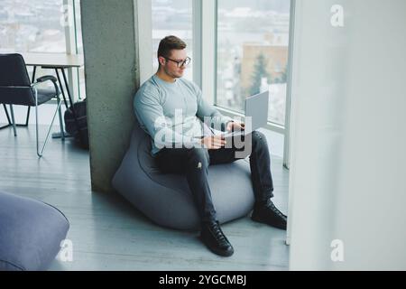 Ein junger Manager in Brille und lässiger Kleidung sitzt auf einem weichen Stuhl und arbeitet an einem Laptop. Arbeitsplatz für Fernarbeit. Stockfoto