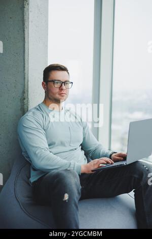 Ein junger Manager in Brille und lässiger Kleidung sitzt auf einem weichen Stuhl und arbeitet an einem Laptop. Arbeitsplatz für Fernarbeit. Stockfoto