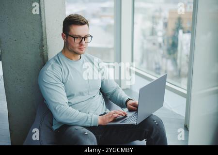 Ein junger Manager in Brille und lässiger Kleidung sitzt auf einem weichen Stuhl und arbeitet an einem Laptop. Arbeitsplatz für Fernarbeit. Stockfoto