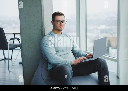 Ein junger Manager in Brille und lässiger Kleidung sitzt auf einem weichen Stuhl und arbeitet an einem Laptop. Arbeitsplatz für Fernarbeit. Stockfoto