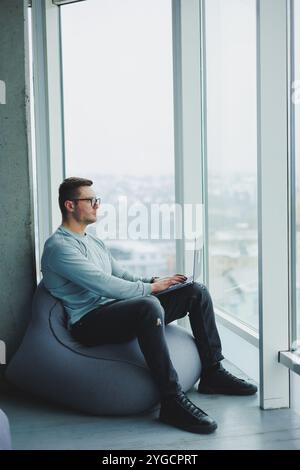 Ein junger Manager in Brille und lässiger Kleidung sitzt auf einem weichen Stuhl und arbeitet an einem Laptop. Arbeitsplatz für Fernarbeit. Stockfoto
