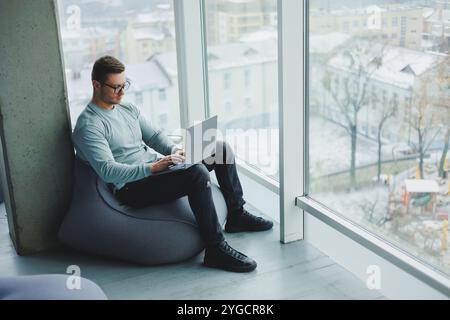Ein junger Manager in Brille und lässiger Kleidung sitzt auf einem weichen Stuhl und arbeitet an einem Laptop. Arbeitsplatz für Fernarbeit. Stockfoto