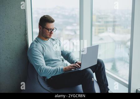 Ein junger Manager in Brille und lässiger Kleidung sitzt auf einem weichen Stuhl und arbeitet an einem Laptop. Arbeitsplatz für Fernarbeit. Stockfoto