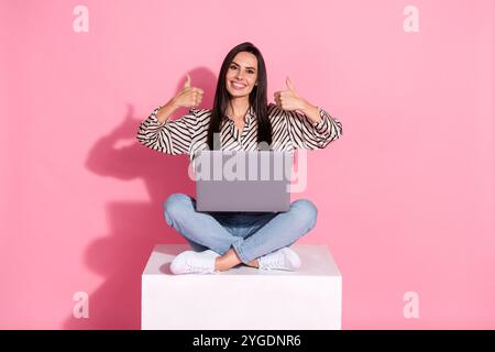Junge Frau in lässigen Jeans und gestreiftem Hemd mit Laptop vor rosa Hintergrund, mit positiver Geste mit zwei Daumen nach oben Stockfoto